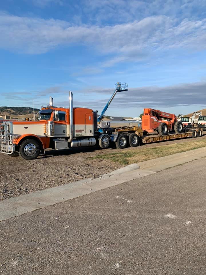 Orange and white semi-truck with flatbed trailer carrying a lift and telehandler. Blue sky in the background.
