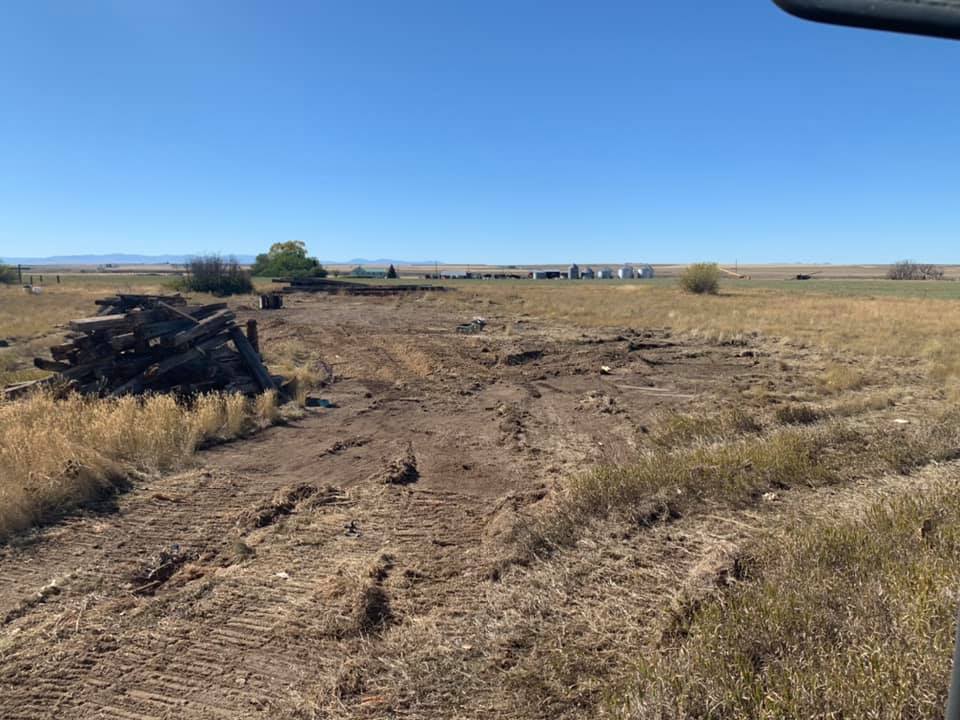 Dirt clearing in a field, with a pile of logs on the left and farm structures in the distance under a blue sky.