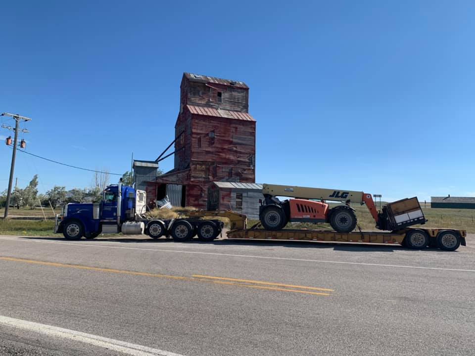 A large truck hauling a dilapidated grain silo and a forklift on a flatbed trailer.
