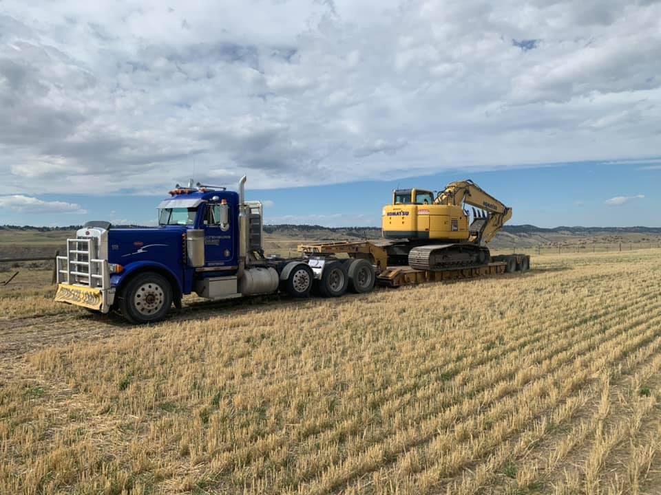 Blue semi-truck transporting a yellow excavator on a flatbed trailer across a harvested field under a cloudy sky.