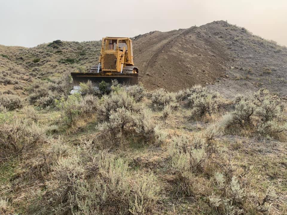 Yellow bulldozer pushing earth on a hillside covered with gray-green shrubs, in a rural setting.