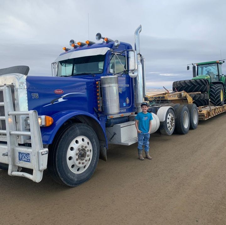 A young person stands beside a blue Peterbilt truck towing a tractor on a flatbed trailer.