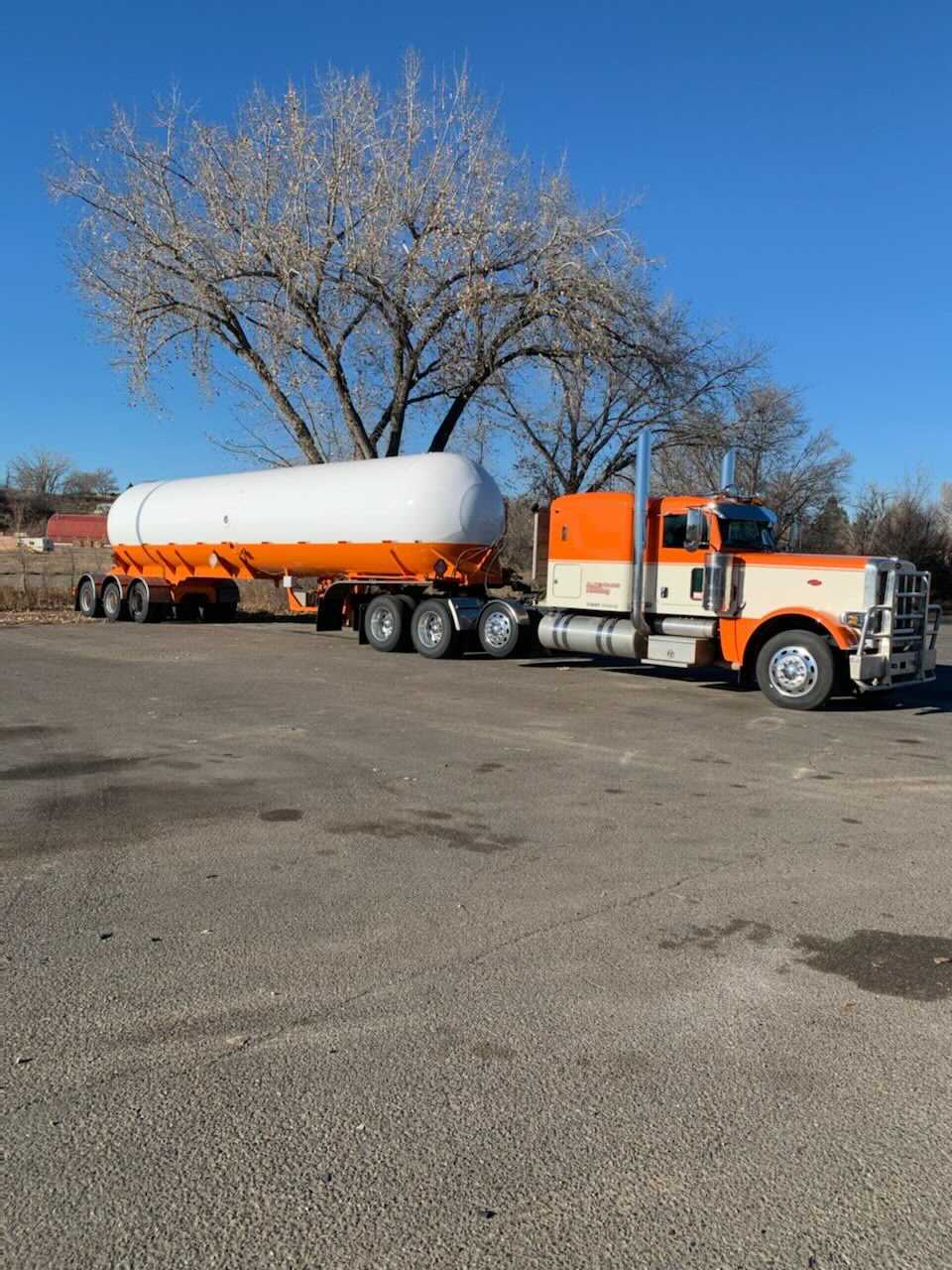 Orange and white tanker truck and trailer parked on pavement in front of a tree against a blue sky.