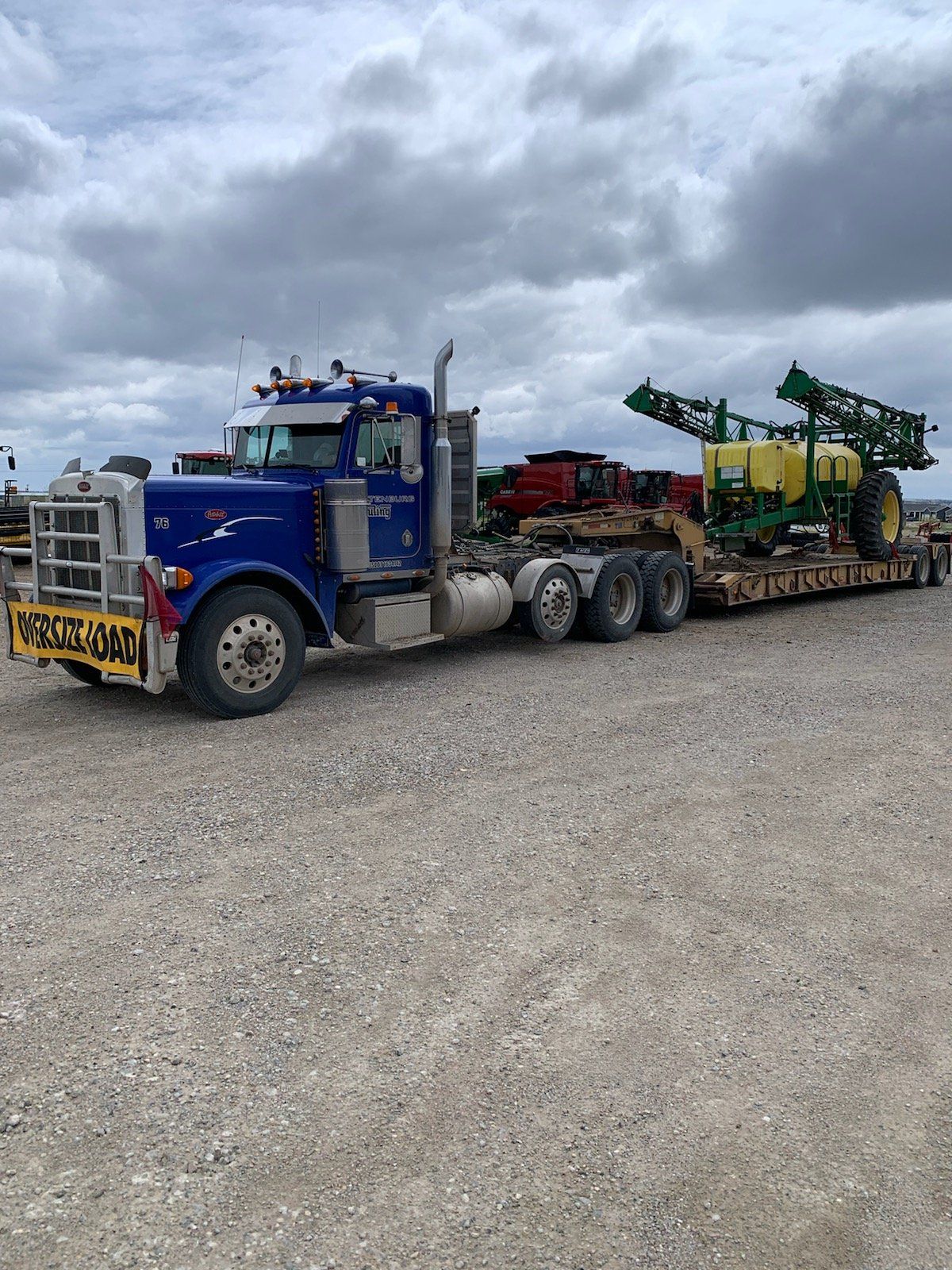 Blue semi-truck with trailer transporting a large green and yellow agricultural sprayer on a gravel lot under a cloudy sky.