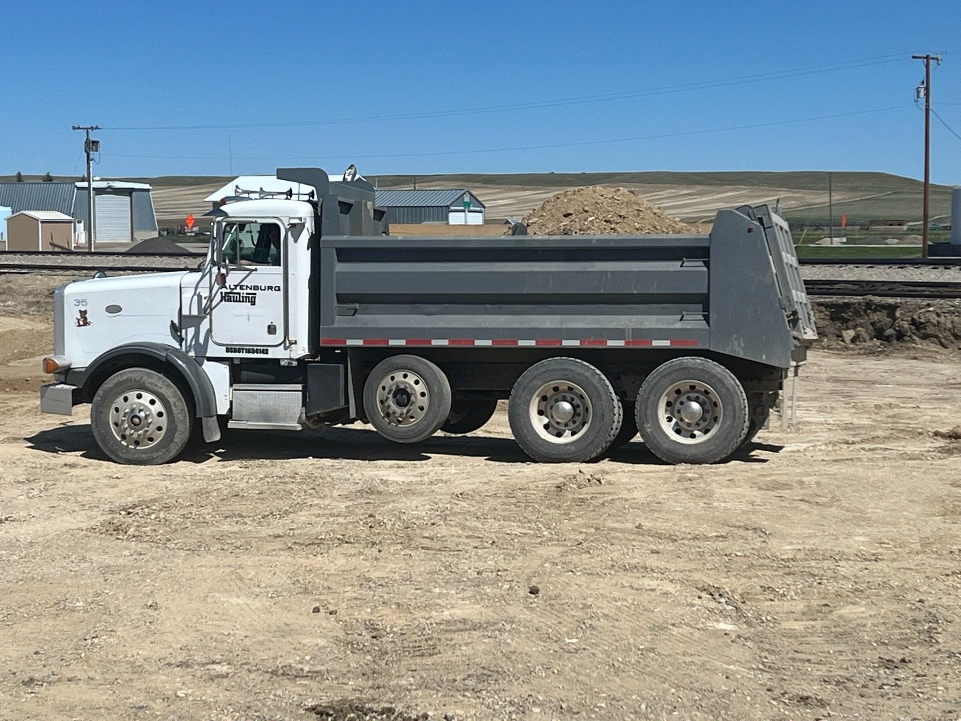 Gray dump truck on dirt, near railroad tracks and buildings on a sunny day.