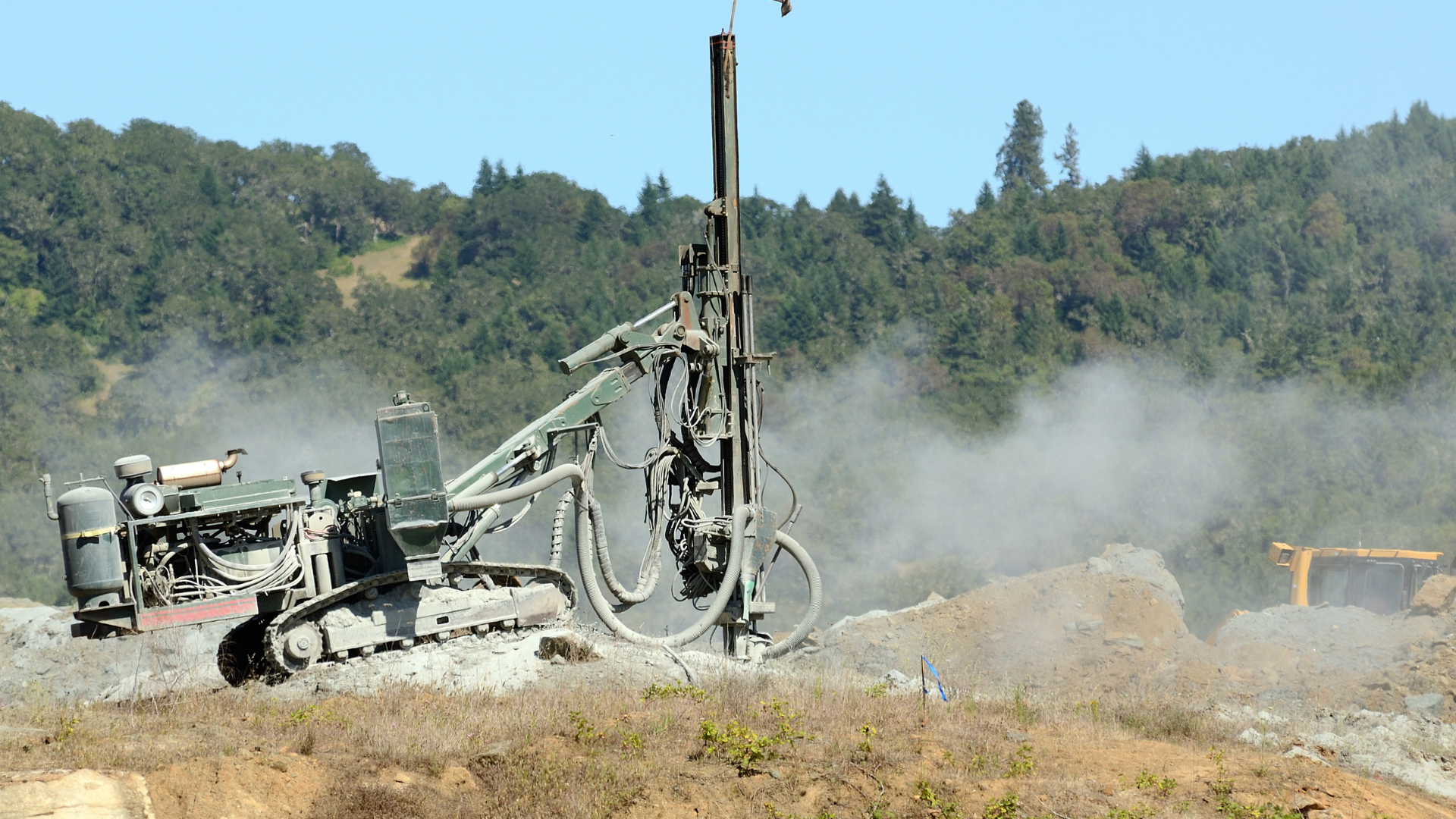 Drilling machine operating on a dusty worksite, trees in the background.