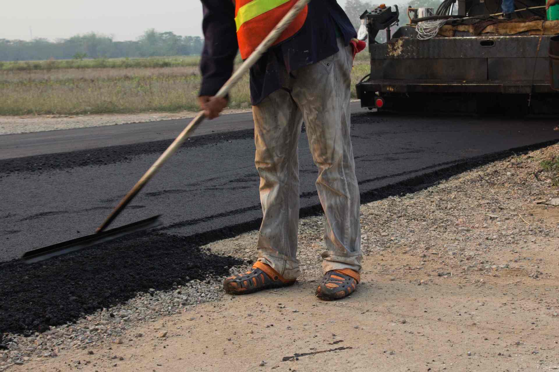 Road worker in orange vest, raking asphalt on a newly paved road.