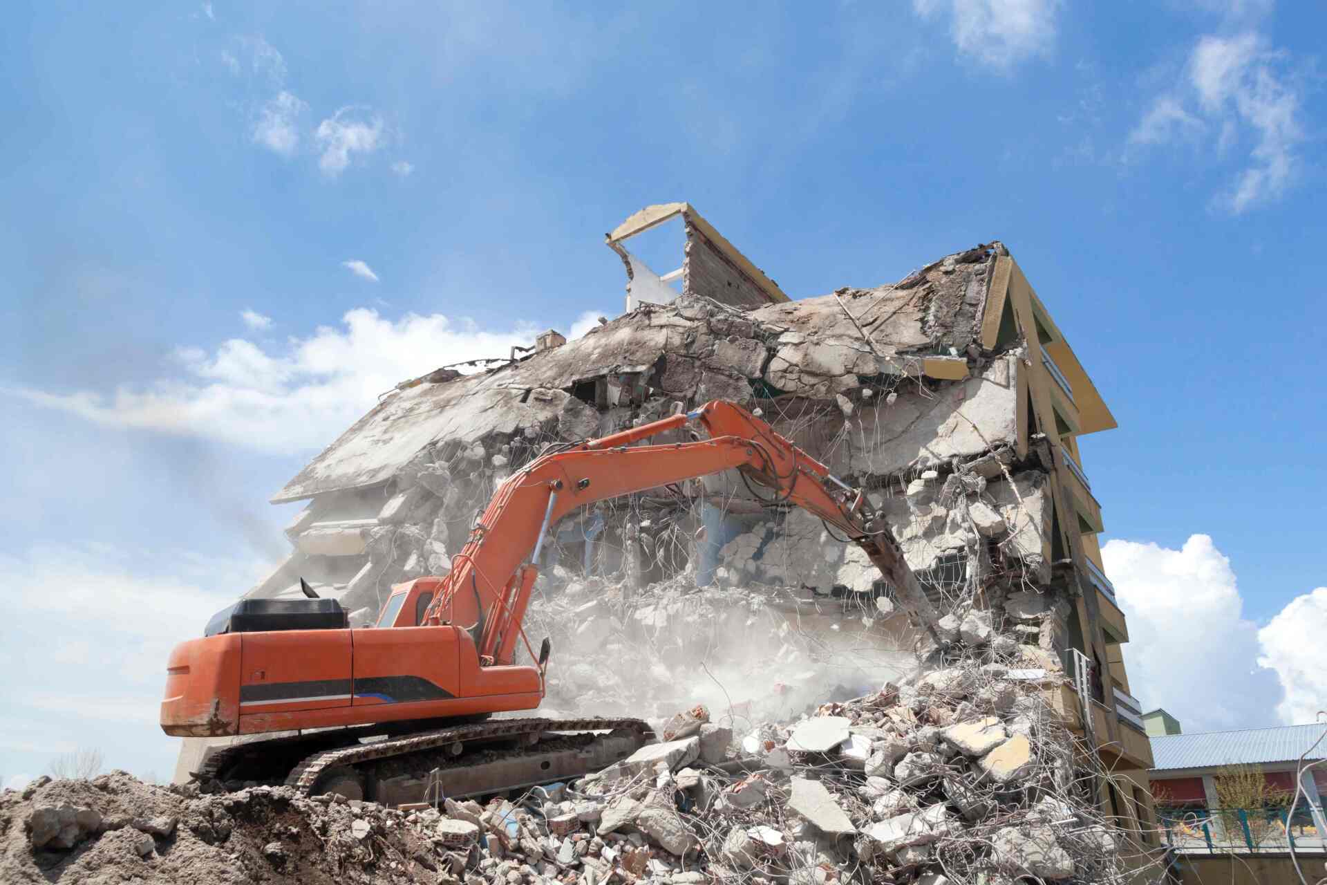 An excavator demolishes a building against a cloudy blue sky.
