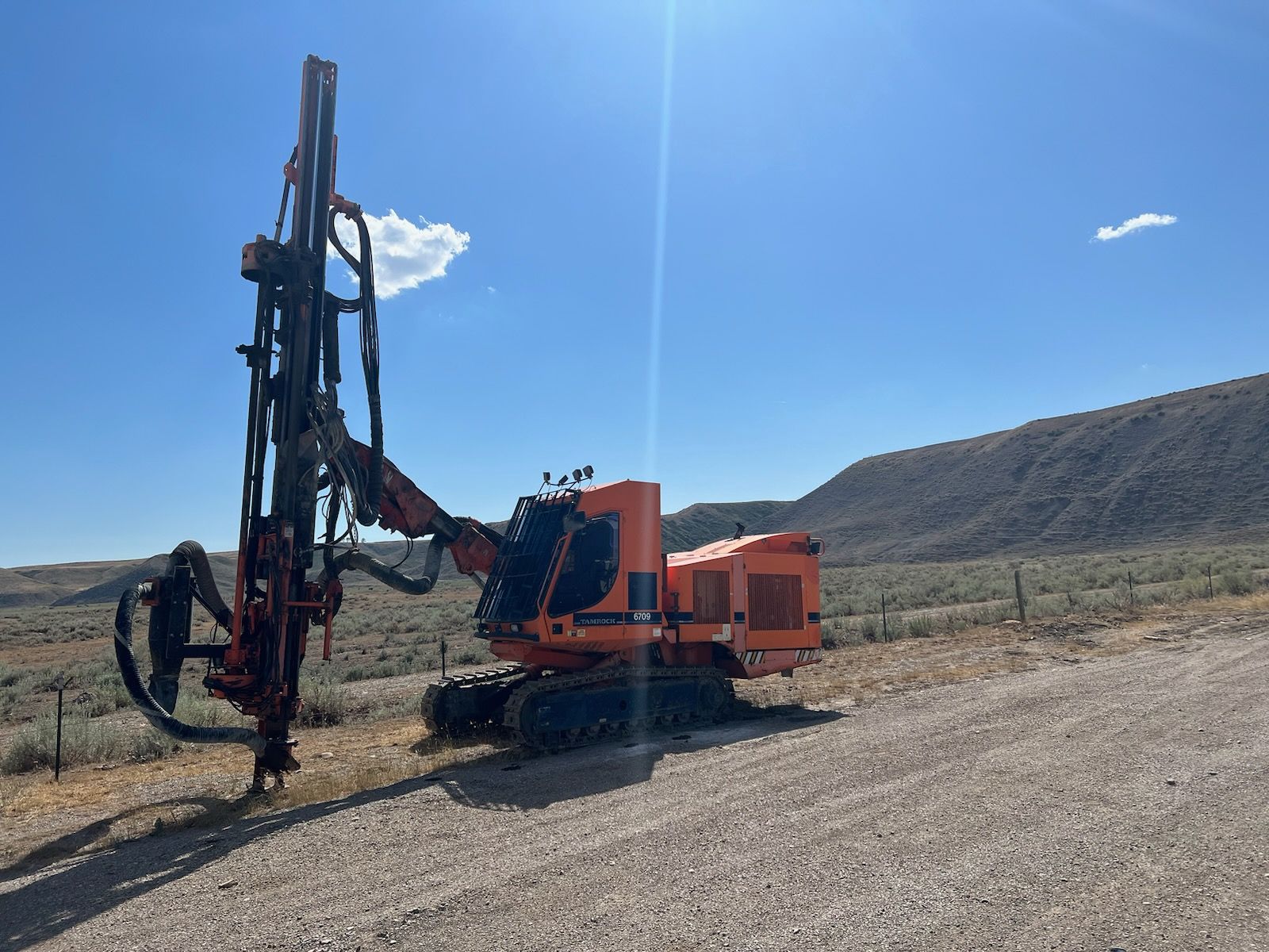 Orange drilling rig on a gravel road, with a hilly landscape and blue sky in the background.