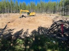 Yellow excavator digs in a brown dirt area surrounded by trees under a blue sky.