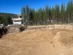 A cleared, level dirt lot with a building in the background and a line of trees against a clear blue sky.