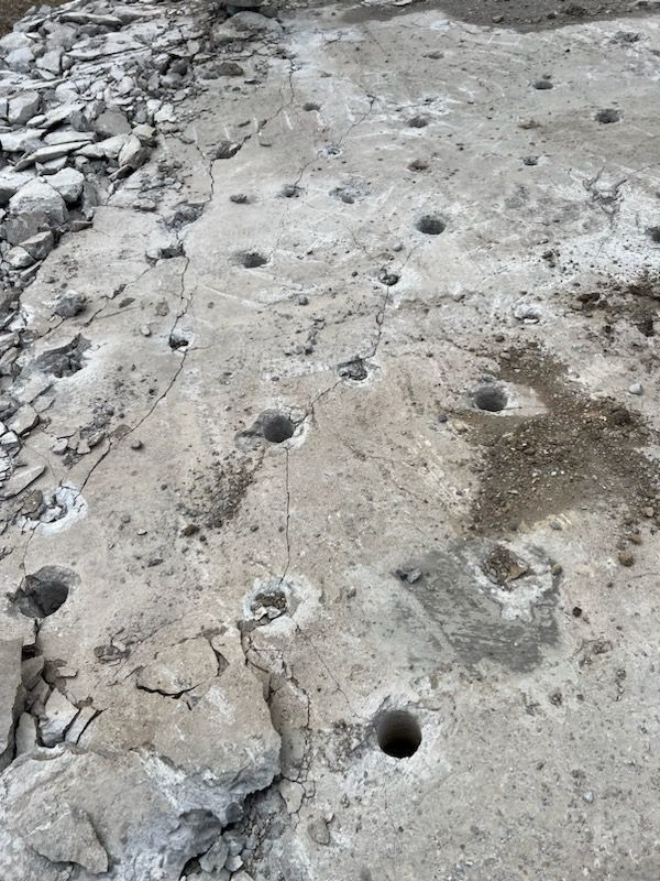 Concrete surface with numerous drilled holes. Debris scattered around the edges, suggesting demolition or construction.