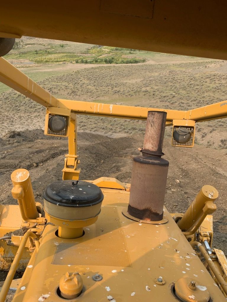 View from inside a yellow bulldozer, overlooking a dirt field and vegetation.