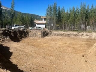 Excavated construction site with a building foundation, mountains, and trees under a blue sky.