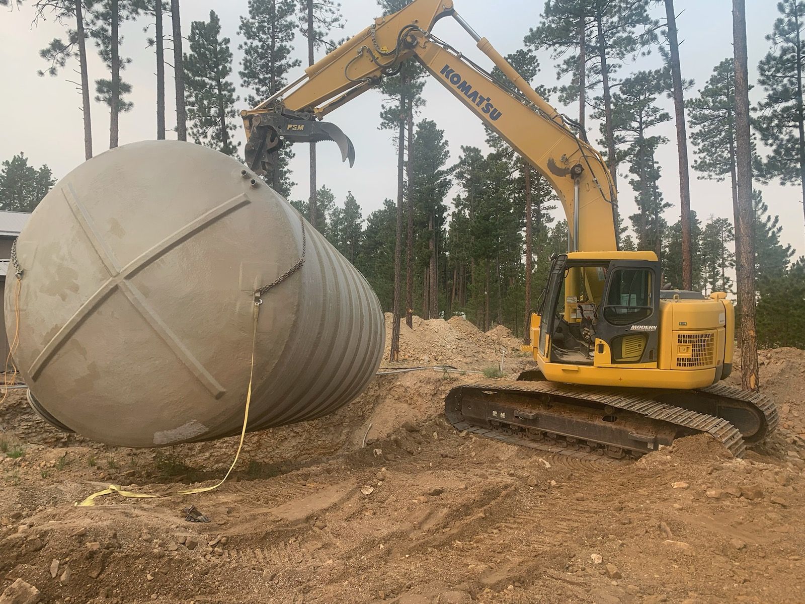 Yellow excavator lifting a large, tan, cylindrical tank in a dirt clearing near trees.