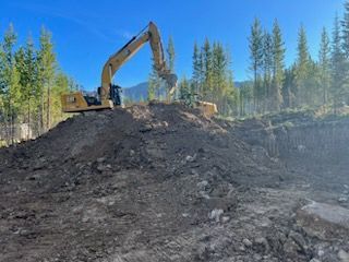 Yellow excavator digging earth on a construction site with trees and mountains in the background under a blue sky.