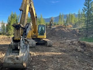 Yellow excavator at work, clearing dirt in a forested area, with another excavator in the background under a blue sky.