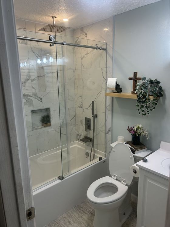 Small white bathroom with a glass shower, marble walls, toilet, sink, and shelf with decor.