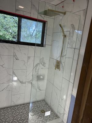 Modern tiled shower with glass partition, rain showerhead, window, and gray patterned floor mat