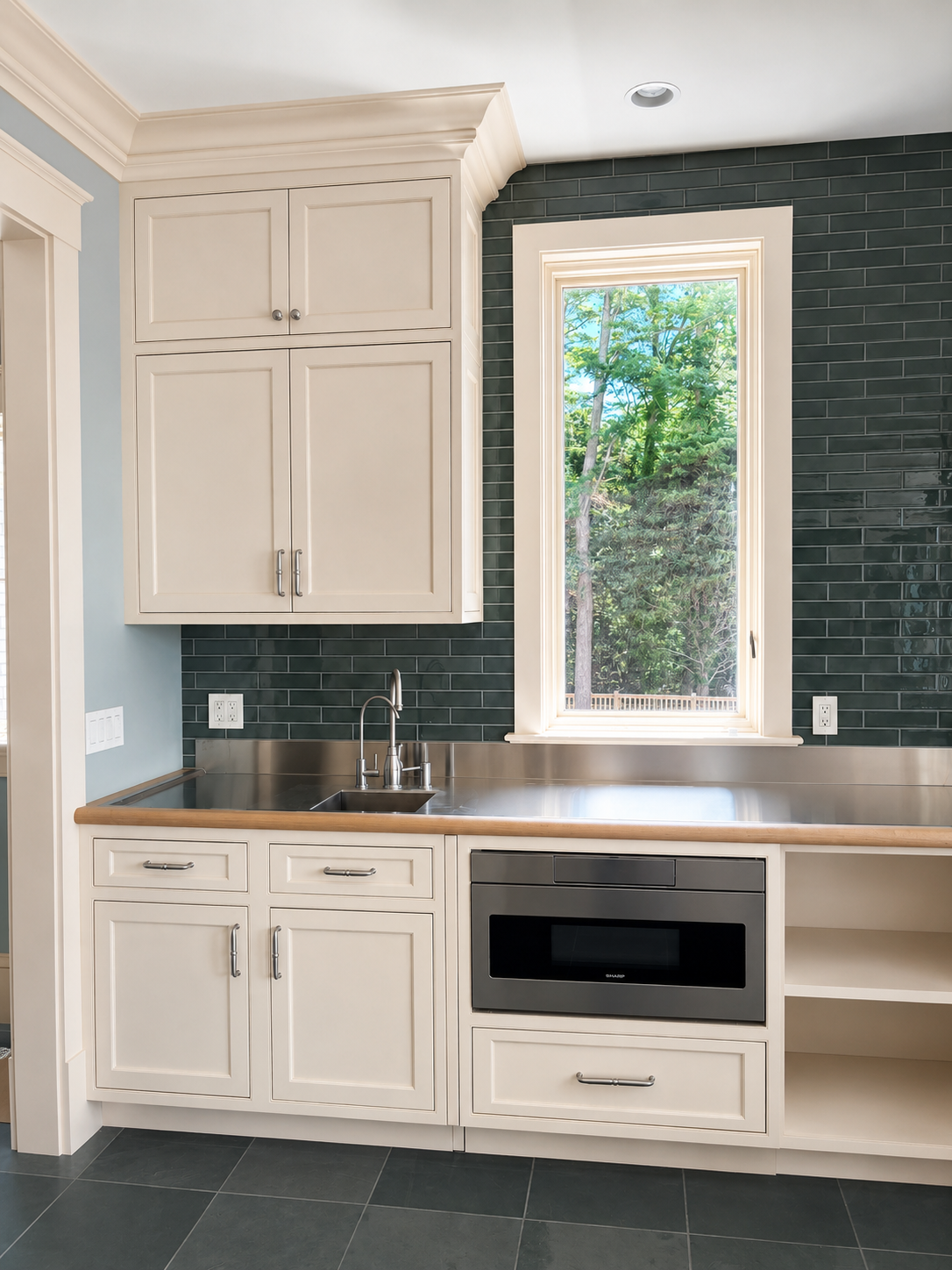 White kitchen with cream cabinets, dark green tile backsplash, wood countertop, and window above the sink.