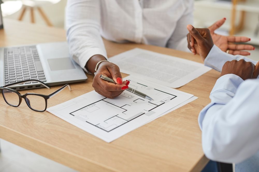 Two people reviewing a floor plan on a desk with a laptop and eyeglasses nearby