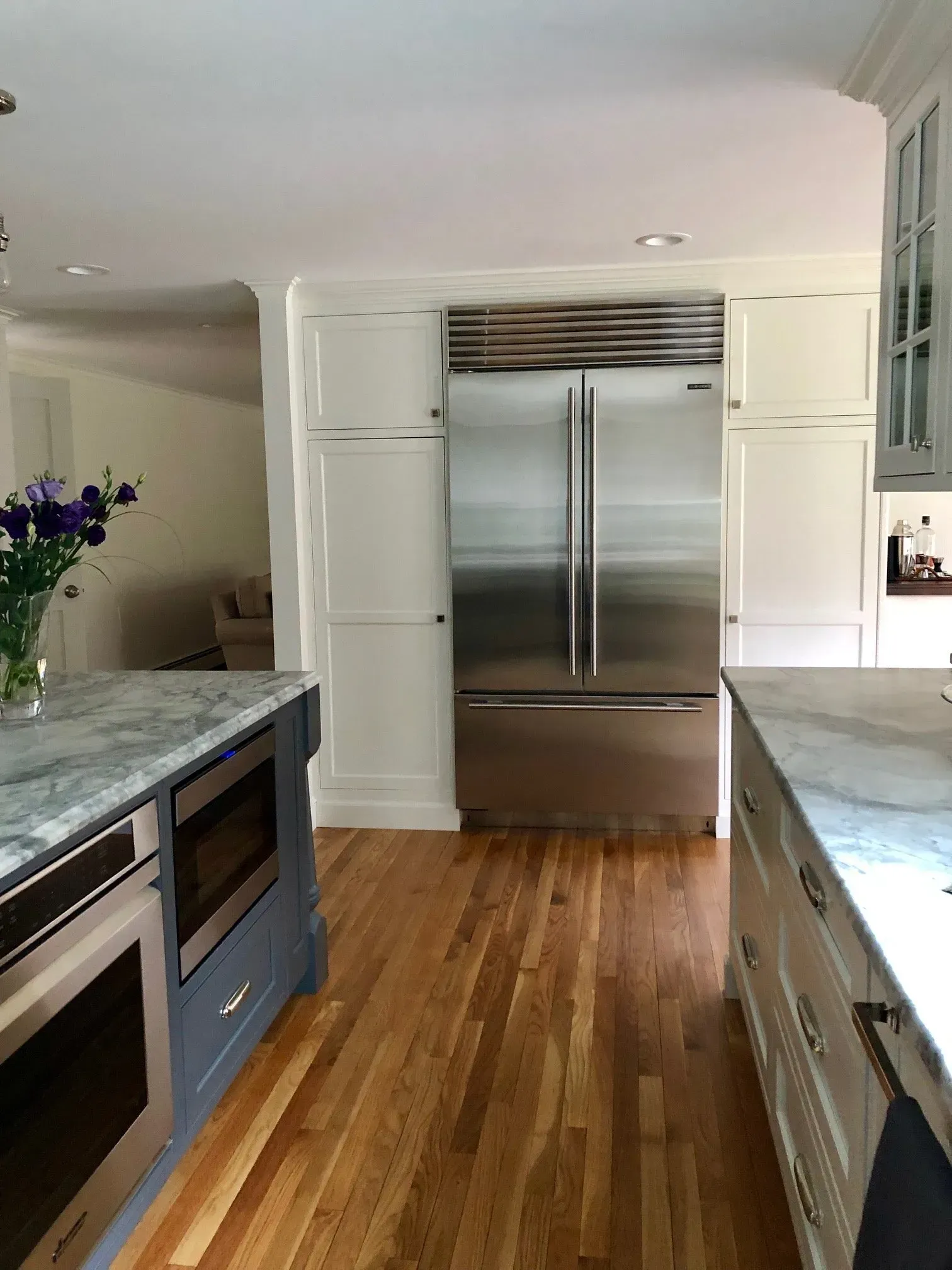 Kitchen with stainless steel refrigerator, white cabinets, blue island, marble countertops, and hardwood floor.