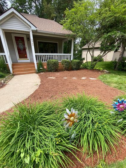 Front yard garden with mulch beds, green shrubs, and a small house porch.