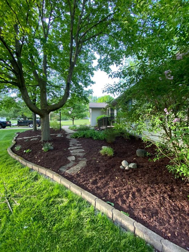 Mulched landscaped yard with mature trees, stone edging, and a small building in the background