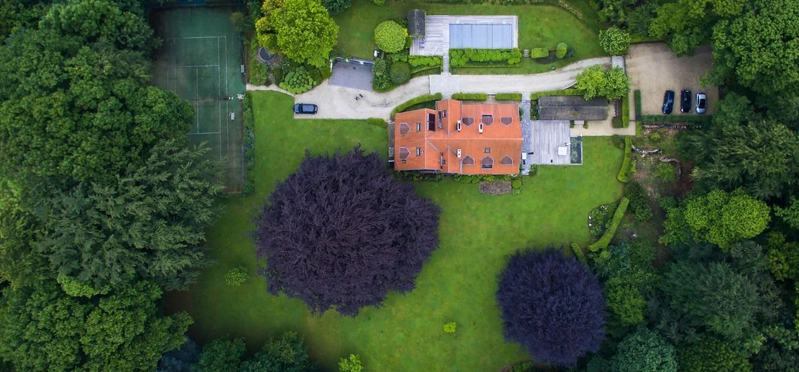 Aerial view of a house with a red roof, solar panels, and a large green lawn surrounded by trees.