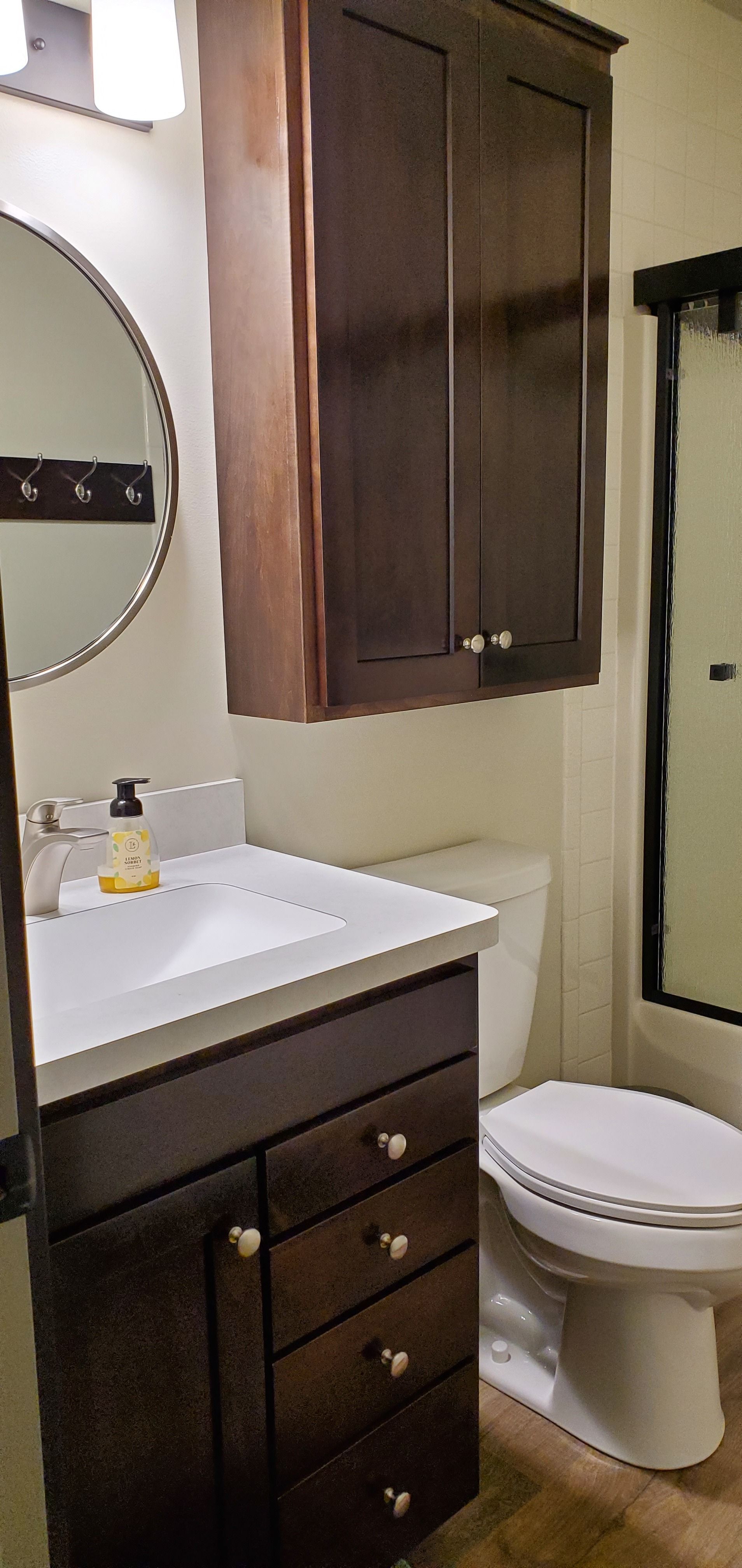 A bathroom featuring a dark wood vanity with drawers, a white sink, a round mirror, and a toilet next to a shower.