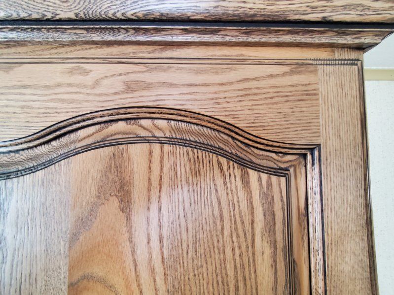 Close-up of a light-brown oak cabinet door with a decorative, arched carved frame and prominent wood grain patterns.