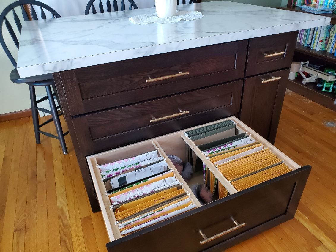 A dark wood kitchen island with a marble top, featuring a bottom drawer pulled open to reveal organized hanging files.