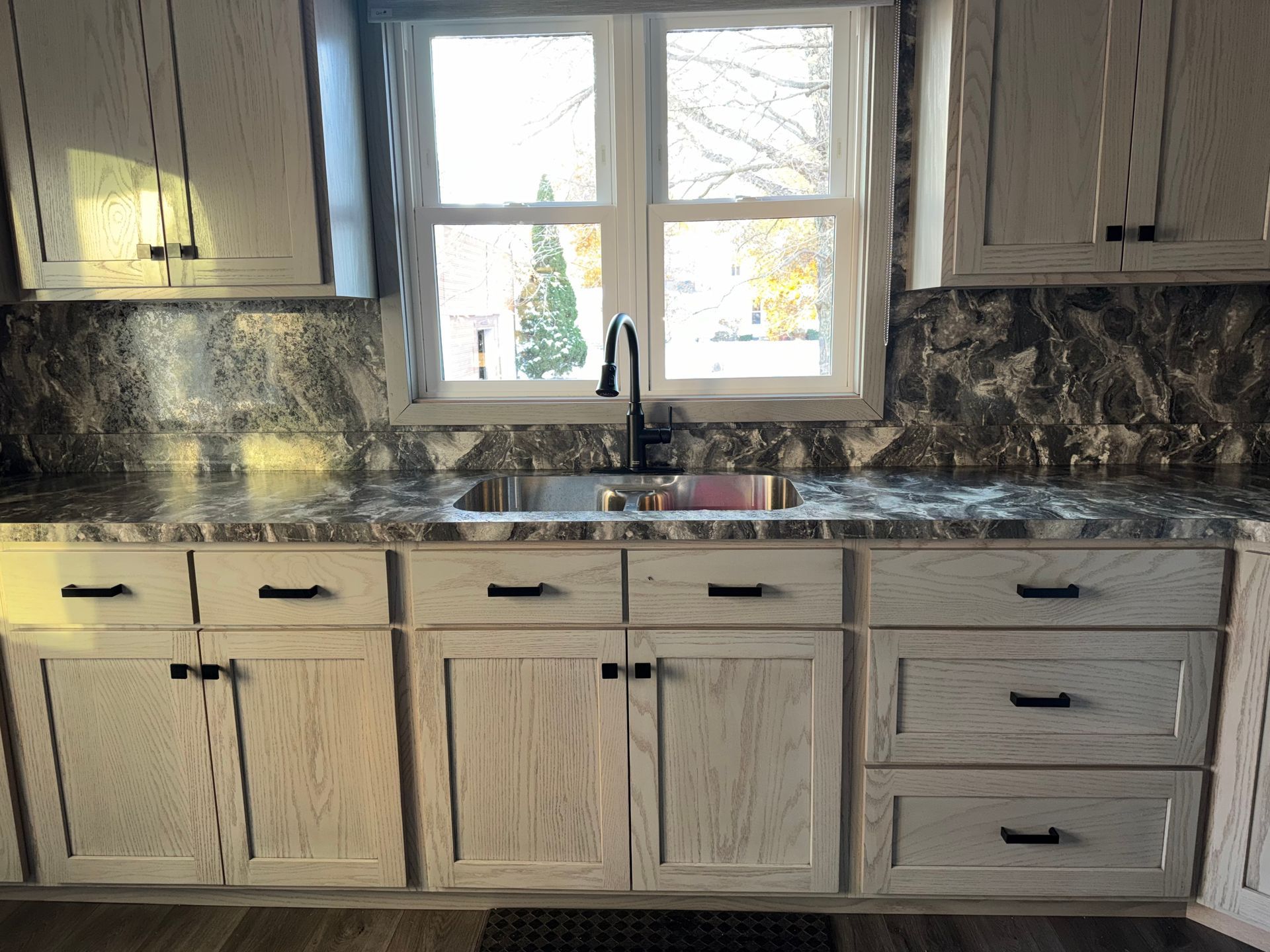 Light-toned wood kitchen cabinets with dark hardware and a marble-patterned countertop under a kitchen window.
