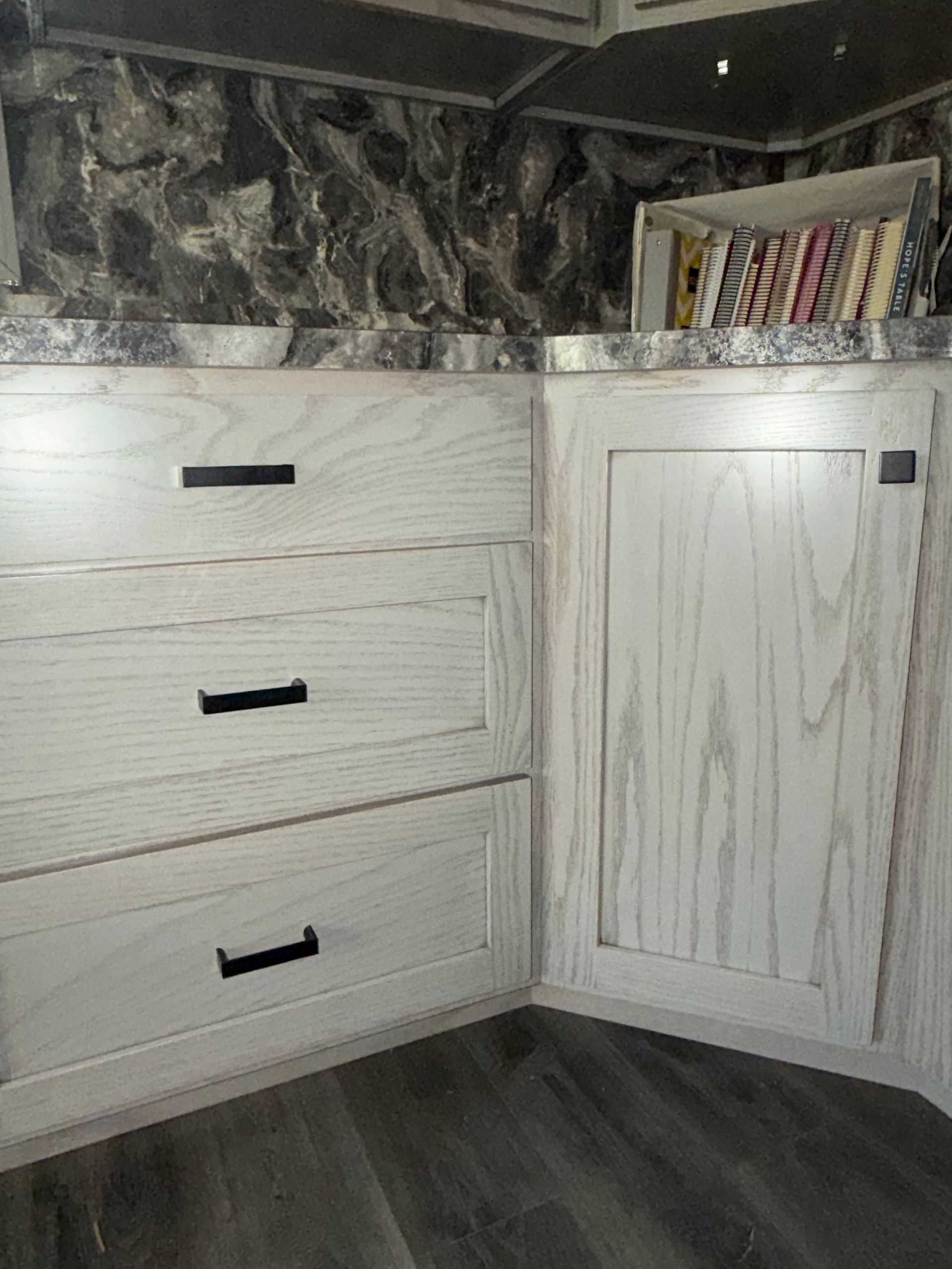 Light wood cabinets with black hardware and a dark countertop in a kitchen corner with a small book shelf.