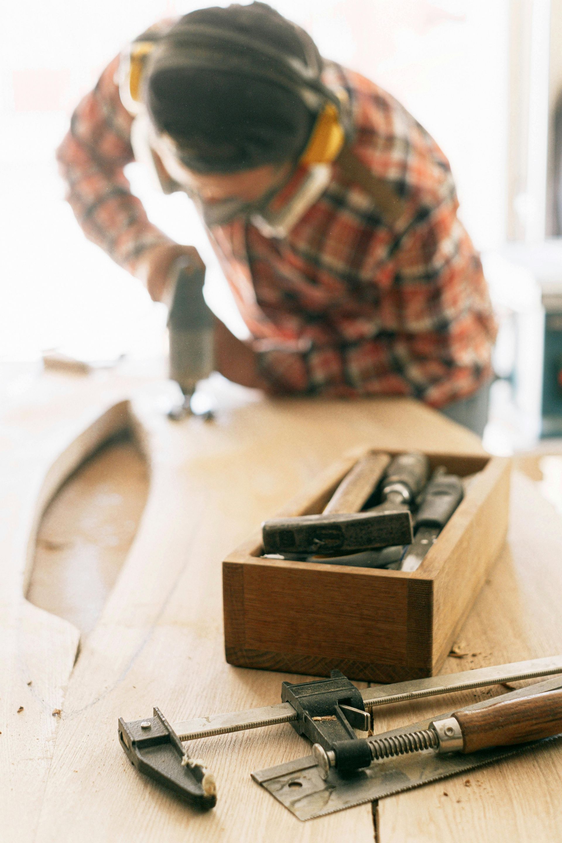 A woodworker wears ear protection while using a power tool on a wooden board, with tools and a clamp on the workbench.
