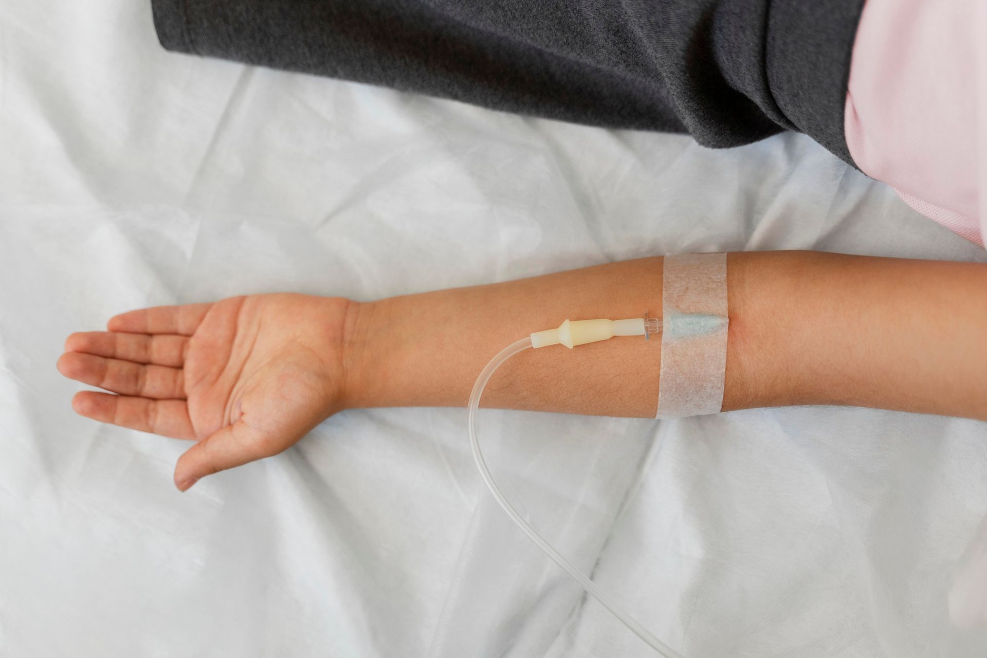 Arm extended on white bed with IV line and transparent medical dressing near the wrist