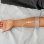 Arm extended on white bed with IV line and transparent medical dressing near the wrist