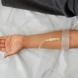 Arm extended on white bed with IV line and transparent medical dressing near the wrist