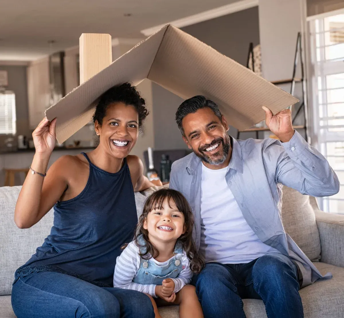 Family sitting on a couch under a cardboard roof, smiling in a bright living room.