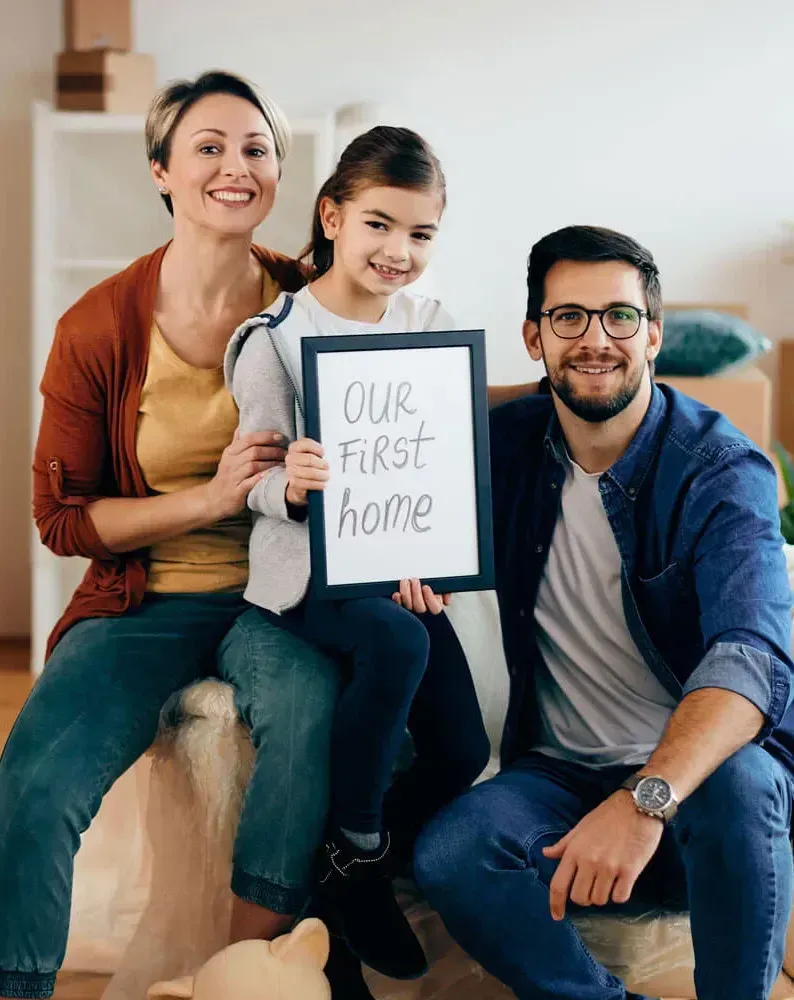Family holding a sign that reads “Our First Home” while sitting together in a living room.