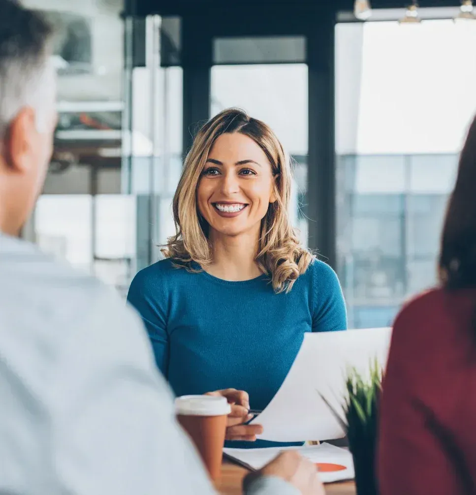 Smiling woman in blue shirt at a meeting table, holding a coffee cup in a bright office