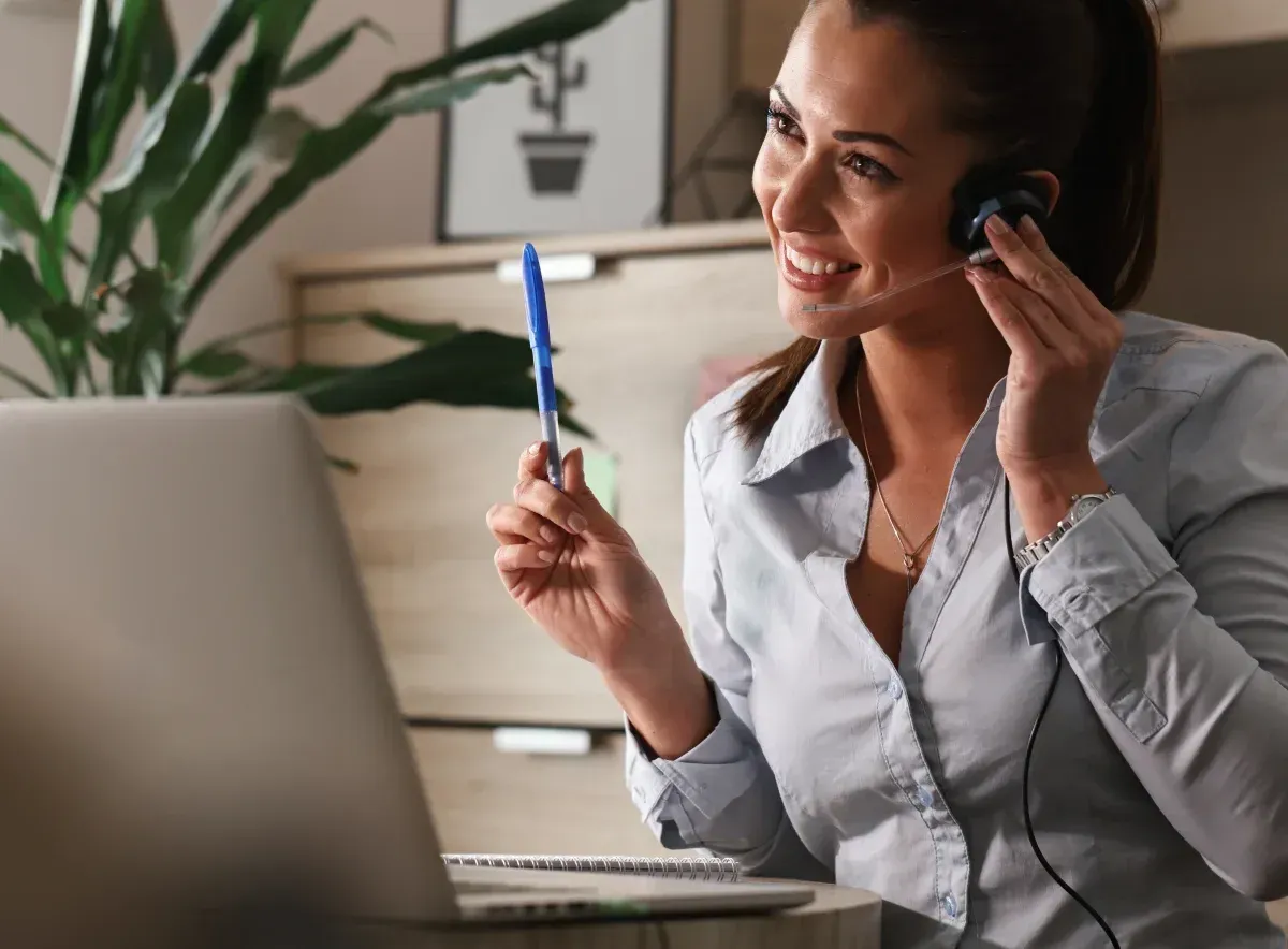 Woman in a white blouse smiling on a headset while holding a pen beside a laptop at her desk