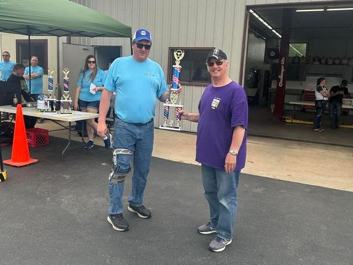 Two men holding a trophy outdoors. One in blue shirt and jeans, one in purple. Other trophies and people in background.