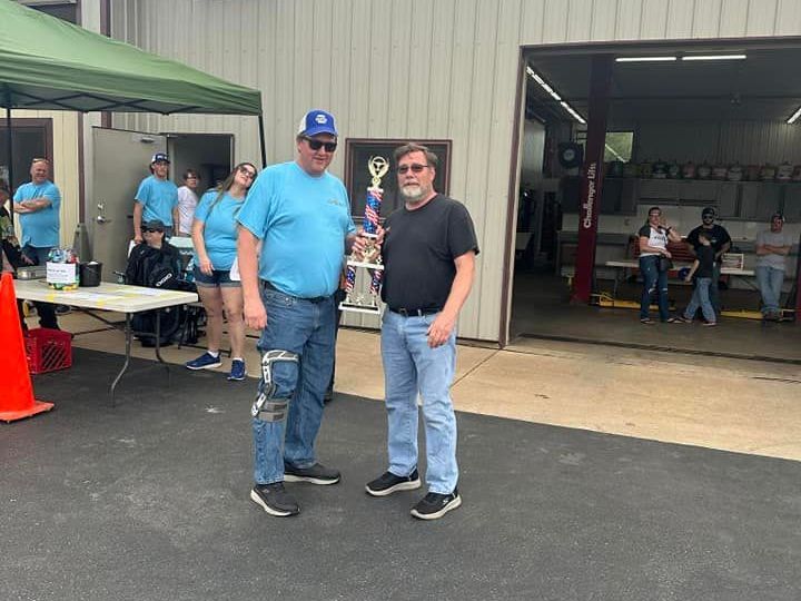 Two men holding a trophy outdoors; one wears a blue shirt and brace, the other a black shirt and jeans.
