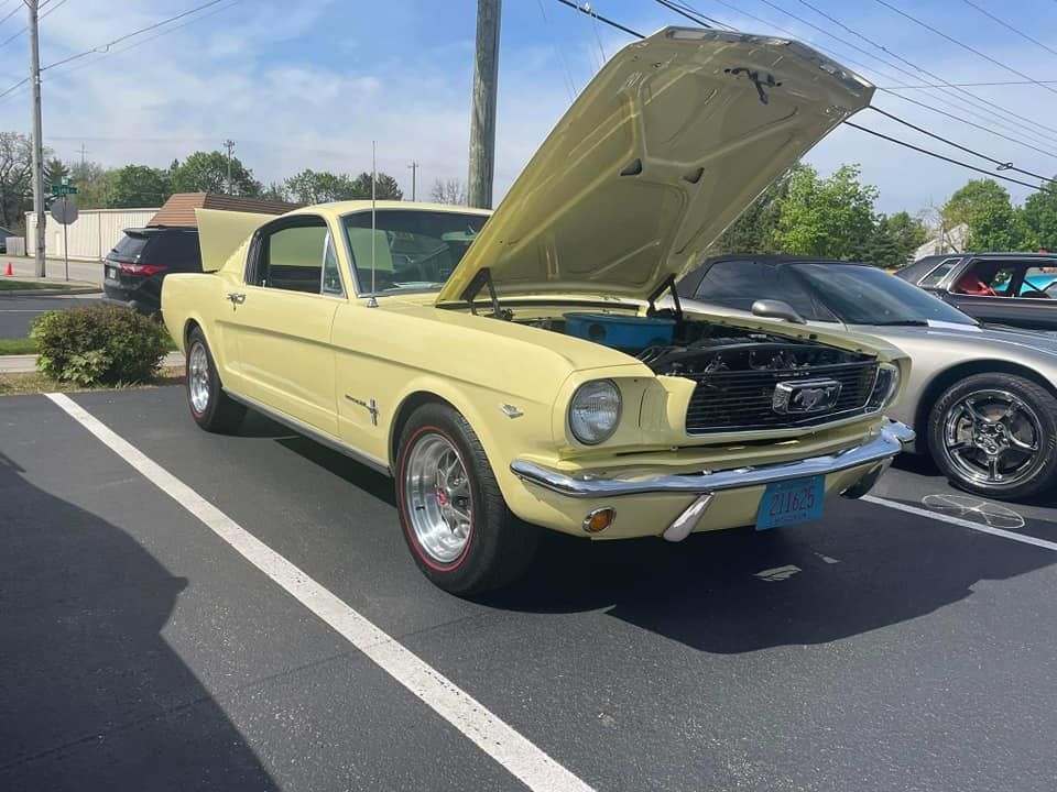 Yellow classic Ford Mustang with hood open parked outside, in a parking lot on a sunny day.