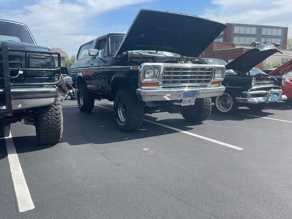 Black Ford Bronco with hood up, parked in a lot with other vintage cars on a sunny day.