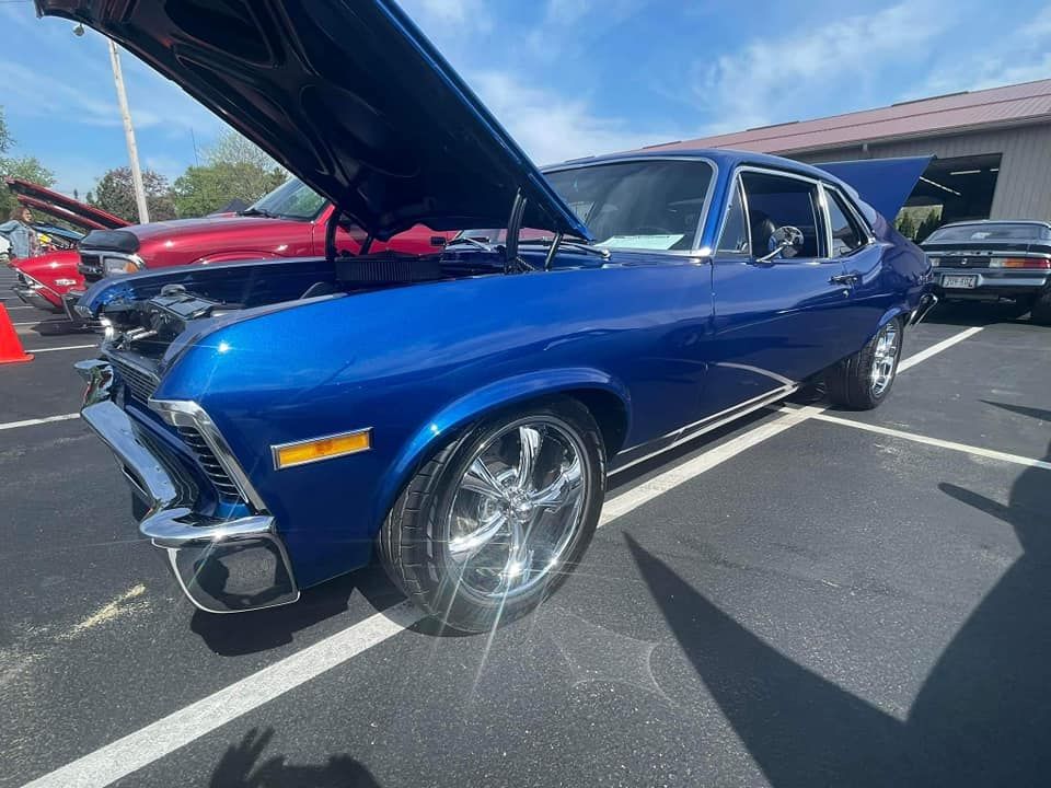 Blue classic Chevy Nova with hood open, parked at a car show on a sunny day.