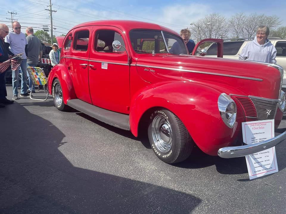 Red classic Ford car at an outdoor car show with onlookers on a sunny day.