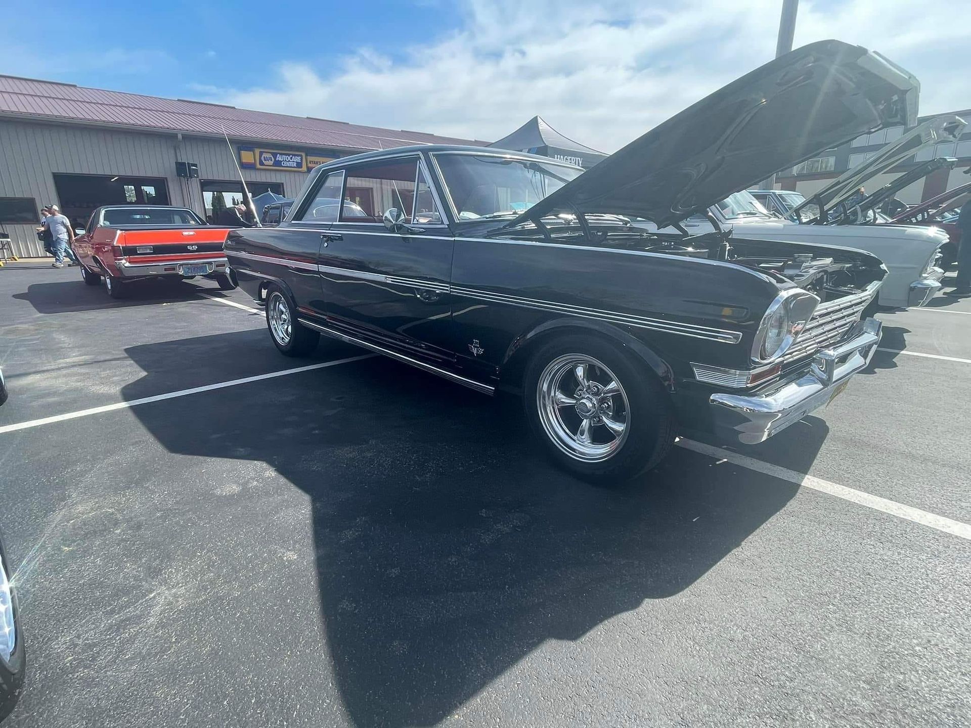 Black classic car with hood up at an outdoor event; other vehicles and building in background.