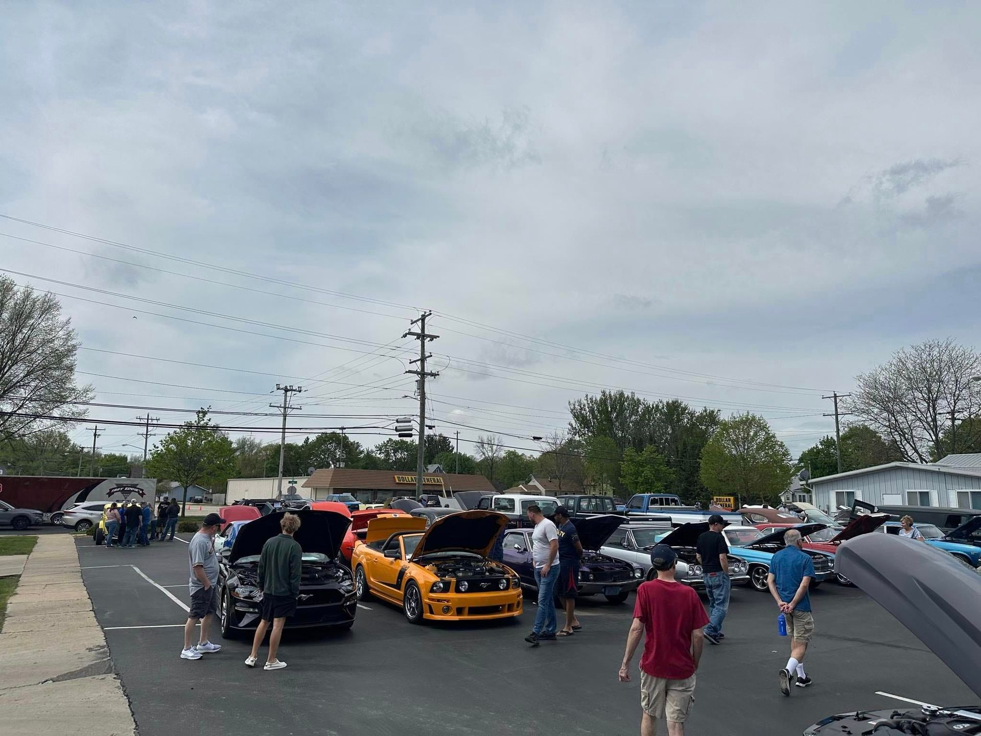 Car show in parking lot; people looking at various cars with open hoods under cloudy sky.